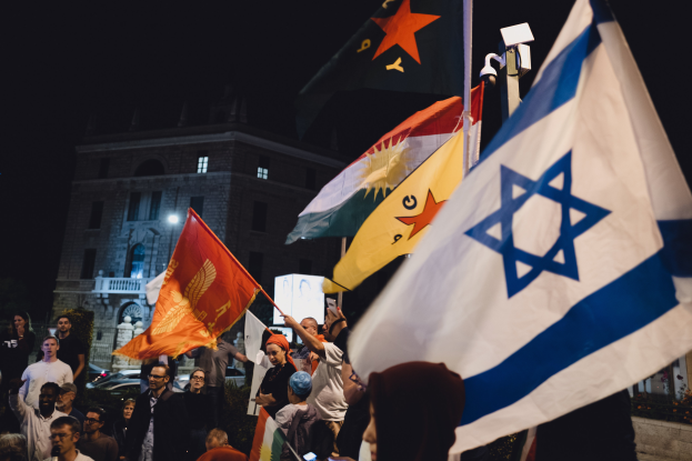 Eine Gruppe von Menschen steht vor einem Gebäude und hält Fahnen während einer anti-israelischen Demonstration bei Nacht, mit Fahrzeugen und Pflanzen im Hintergrund.
