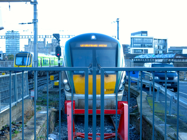 Zwei Züge auf dem Gleis mit einem Zaun im Vordergrund, Masten auf dem Land, einer mit einer Signalleuchte, ein Auto auf der Straße rechts, Gebäude im Hintergrund und der Himmel darüber.