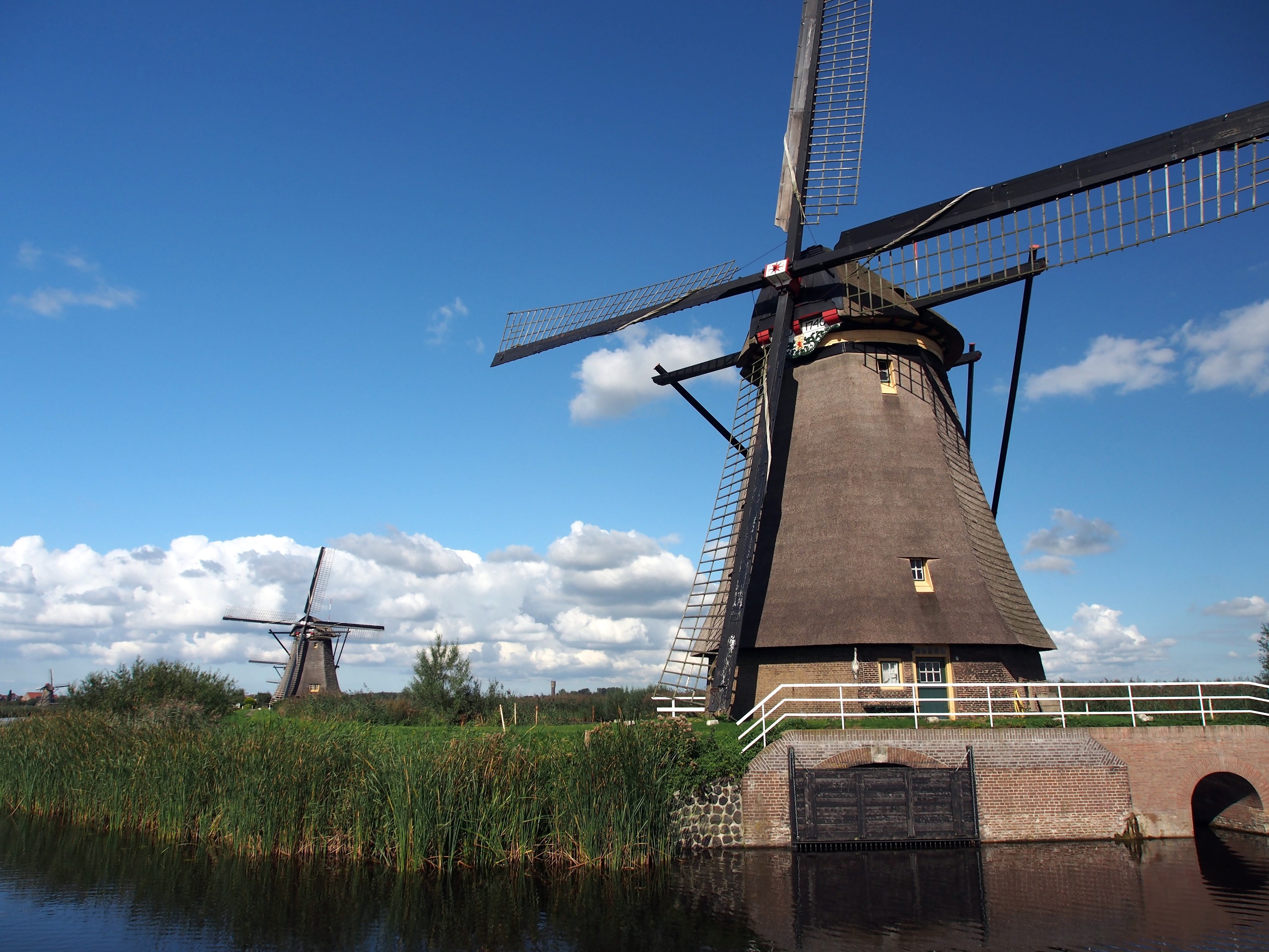 Windmühle in Kinderdijk, Niederlande, umgeben von Wasser, Gras, einer Brücke, Geländern, Bäumen und einem bewölkten Himmel.