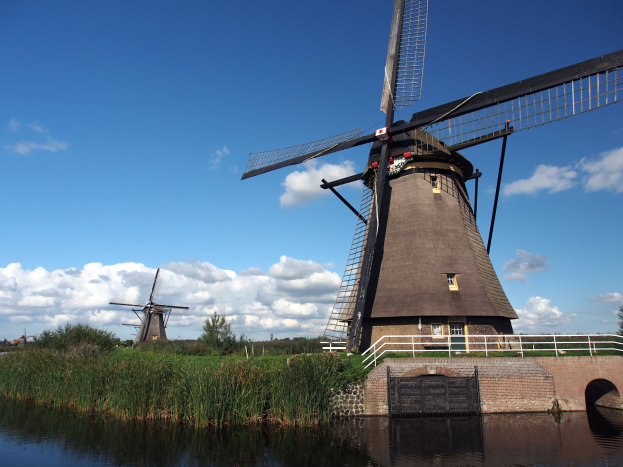 Windmühle in Kinderdijk, Niederlande, umgeben von Wasser, Gras, einer Brücke, Geländern, Bäumen und einem bewölkten Himmel.
