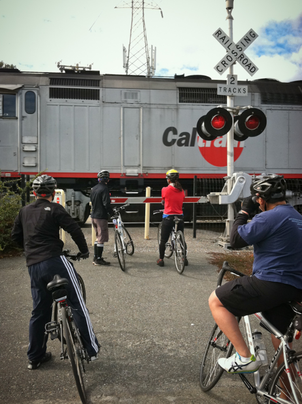 Vier Personen mit Fahrrädern stehen auf einer Straße neben einem Bahnsignal und einem Tor.