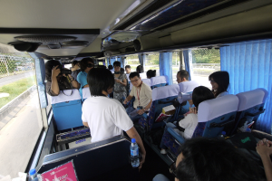 Ein Bus voller Passagiere auf einer Straße, mit einem Zaun auf der linken Seite, einer Straßenabsperrung auf der rechten Seite und Bäumen und Pflanzen im Hintergrund; eine Person hält eine Kamera, und Lautsprecher sind auf dem Dach eines Fahrzeugs angebracht.