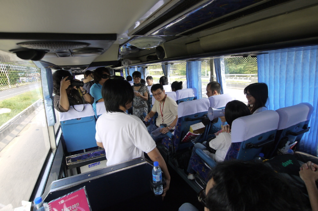 Ein Bus voller Passagiere auf einer Straße, mit einem Zaun auf der linken Seite, einer Straßenabsperrung auf der rechten Seite und Bäumen und Pflanzen im Hintergrund; eine Person hält eine Kamera, und Lautsprecher sind auf dem Dach eines Fahrzeugs angebracht.