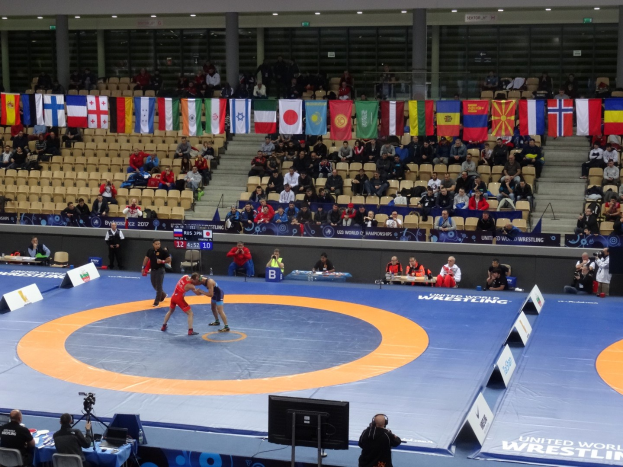 Zwei Wrestler kämpfen in der Mitte einer großen Arena, umgeben von sitzenden und stehenden Zuschauern, mit Kameras, Monitoren und Flaggen im Hintergrund.