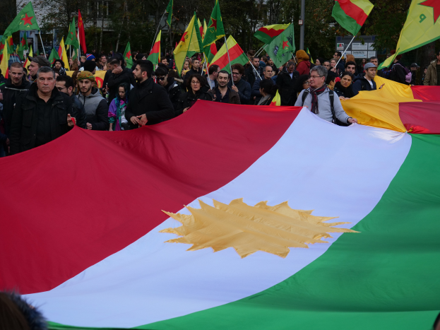 Eine große Gruppe von Menschen mit Fahnen vor einem Gebäude, mit Bäumen und Pfählen im Hintergrund, die wahrscheinlich an einer Protestaktion in Kurdistan teilnehmen.
