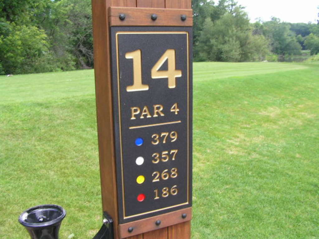 Ein hölzerner Pfahl mit Zahlen darauf steht vor einem grasbewachsenen Bereich mit Bäumen.