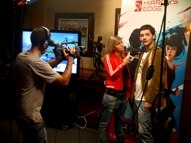 Ein Mann in einem Headset hält eine Videokamera und filmt einen Mann und eine Frau, die ein Interview führen, während im Hintergrund ein Fernseher und ein Foto Rahmen zu sehen sind.