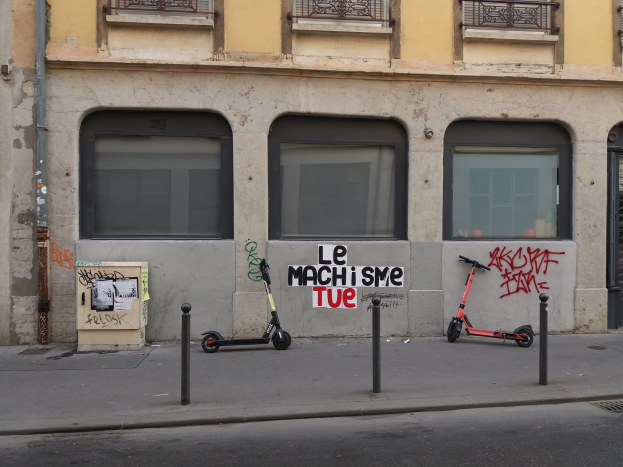 Zwei Roller auf der Straßenseite neben einem Geb"aude mit Fenstern, Gel"andern und einem Rohr geparkt, mit Masten und einer Tafel mit Text in der N"ahe.