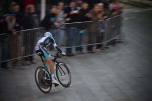 Eine Person fährt ein Fahrrad bei einem Rennen, mit Zuschauern hinter einem Zaun zur linken Seite.