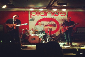 Drei Männer auf der Bühne bei einem Konzert: einer spielt Schlagzeug in der Mitte, und zwei spielen Gitarre und singen auf der linken und rechten Seite, mit einem Banner an der Wand und Lampen, die von der Decke hängen.