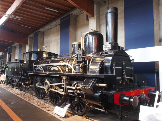 Eine gro├če, alte Dampflokomotive wird in einem Museums Setting gezeigt, positioniert auf Eisenbahnschienen mit Informationsschildern, St├Ąnden und anderen Ausstellungen in der N├Ąhe, vor einer Wand mit ├ťberkopfbeleuchtung.
