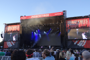 Eine große Menschenmenge steht vor einer Bühne auf dem Shaky Knees Music Festival, wo mehrere Musiker unter hellen Scheinwerfern und Bannern spielen, mit einem Geländer unten und einem klaren blauen Himmel im Hintergrund.