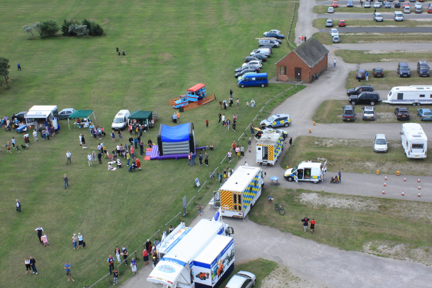 Eine Vogelperspektive eines Parkplatzes mit vielen Fahrzeugen, einigen Menschen in der Nähe von Baracken, einer zentralen Hütte und einigen Bäumen auf der linken Seite.