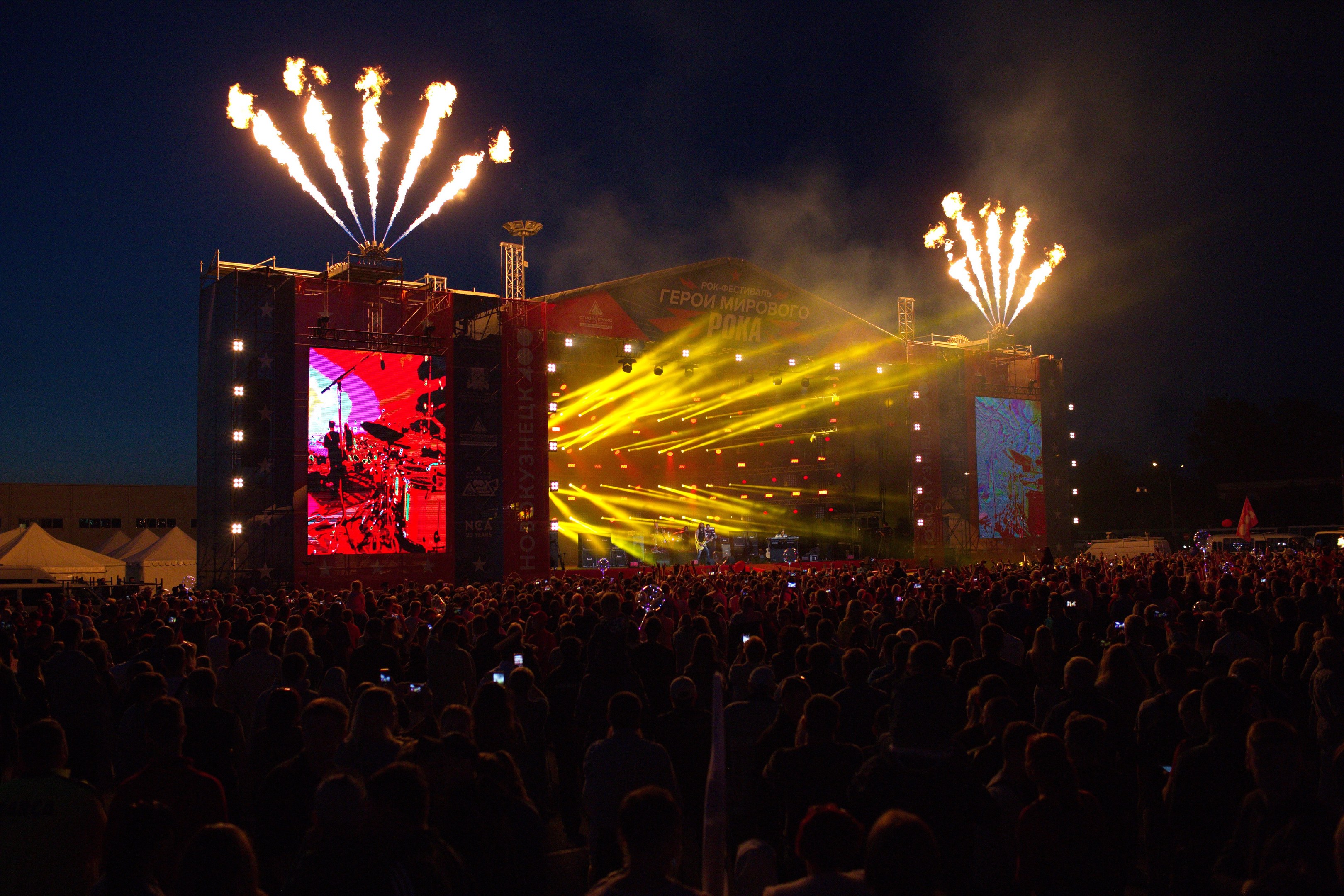 Große Menschenmenge auf einem Musikfestival mit beleuchteter Bühne und Feuerwerk am dunklen Himmel.