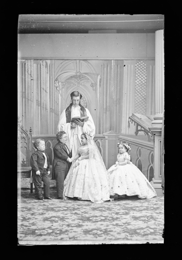 Ein altes Schwarz-Weiß-Foto einer Braut in einem weißen Kleid und Schleier, eines Bräutigams mit einem Buch in der Hand und ihren Kindern, die vor ihnen stehen, mit Säulen und einer Wand im Hintergrund.
