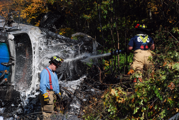 Zwei Feuerwehrmänner in Helmen und Handschuhen, einer sprüht Wasser aus einer Leitung auf ein umgestürztes Auto im Wald, mit Bäumen und Maschendrahtzaun im Hintergrund.