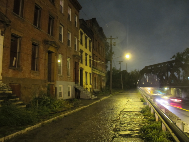 Eine Stadtansicht mit Gebäuden, Treppen mit einem Metallzaun, Strommasten, Laternenmasten mit Kabeln, einer Straße, einem Metallzaun, einer Brücke, Autos, die darunter fahren, und Laternenmasten auf dem Gehweg.