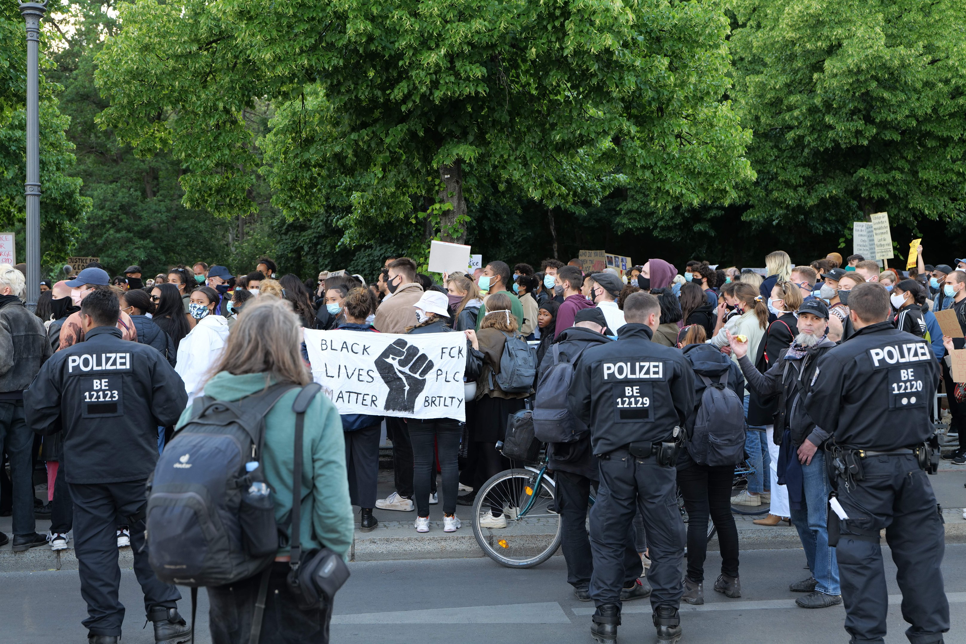 Eine große Gruppe von Menschen steht auf der Seite einer Straße während einer Black Lives Matter Demonstration in Berlin, einige halten Schilder und andere tragen Mützen und Taschen, mit einem Fahrrad im Vordergrund und Bäumen und einem Pfahl im Hintergrund.