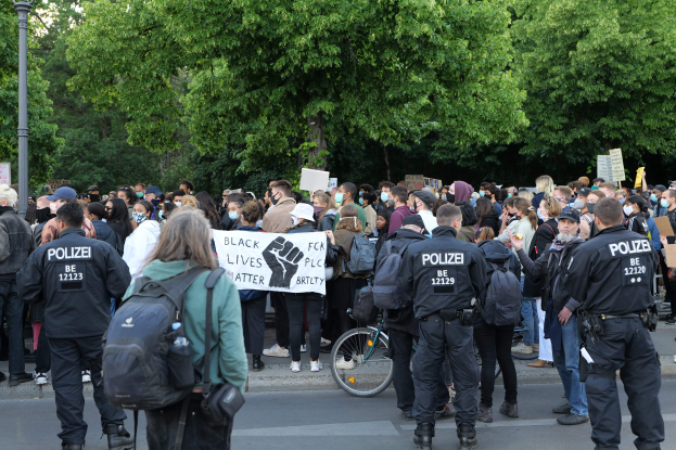 Eine große Gruppe von Menschen steht auf der Seite einer Straße während einer Black Lives Matter Demonstration in Berlin, einige halten Schilder und andere tragen Mützen und Taschen, mit einem Fahrrad im Vordergrund und Bäumen und einem Pfahl im Hintergrund.