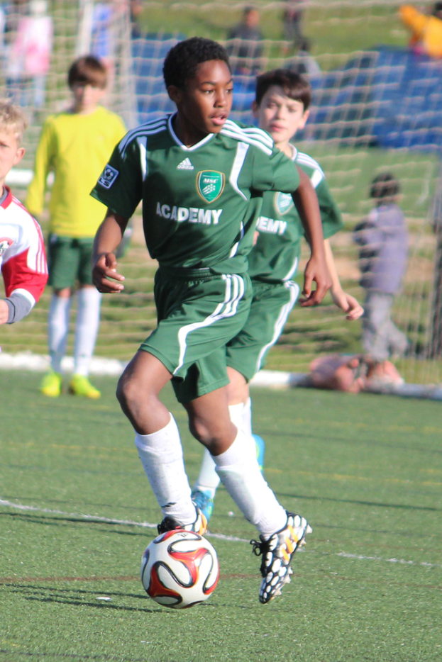 Zwei Personen spielen Fußball in einem Stadion mit wenigen Zuschauern und einem Netz im Hintergrund, ein Ball ist im Spiel.
