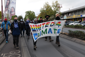 Eine Gruppe von Menschen geht eine Straße entlang, hält eine 'United 4 Climate'-Schlagzeile, trägt Masken und trägt Taschen, mit einem Gehweg, Geländer, Schlagzeilen, Bäumen, Laternen, Fahrzeugen, Gebäuden und einem klaren blauen Himmel im Hintergrund.