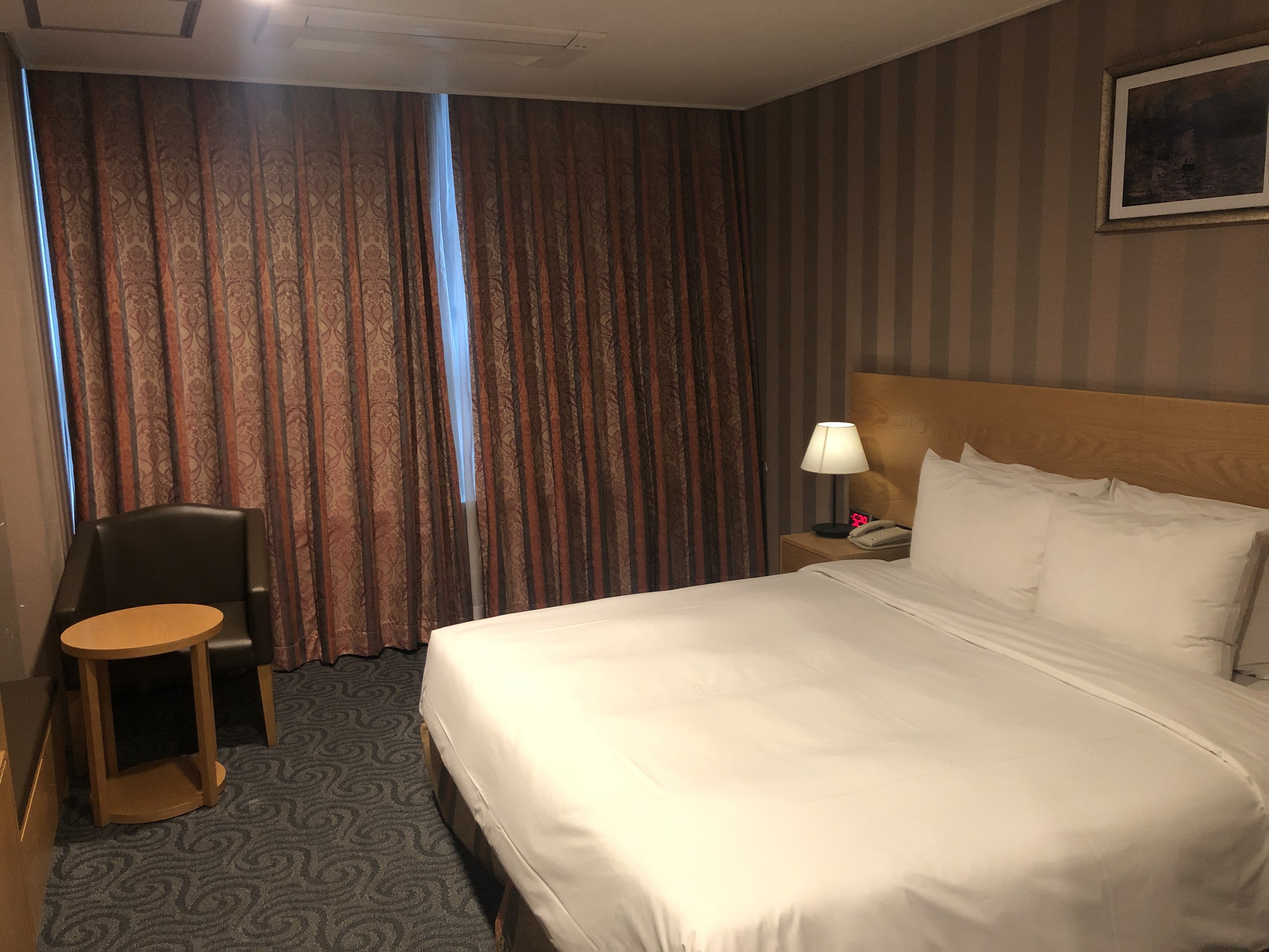 Hotelzimmer mit großem Bett, Stuhl, Tisch, Telefon, Lampe, Foto auf dem Nachttisch, Fenster mit Vorhängen und Deckenbeleuchtung.