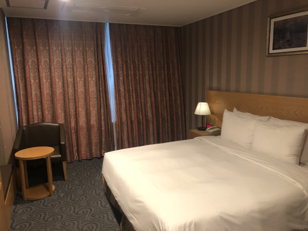 Hotelzimmer mit großem Bett, Stuhl, Tisch, Telefon, Lampe, Foto auf dem Nachttisch, Fenster mit Vorhängen und Deckenbeleuchtung.