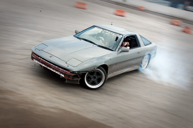 Eine Person mit Helm fährt ein Auto mit Rauch auf einer von orangefarbenen Kegeln umgebenen Strecke, mit einem unscharfen Hintergrund, der hohe Geschwindigkeit andeutet.