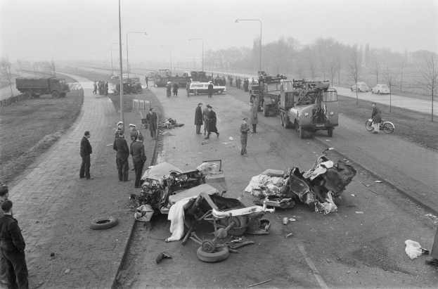 Schwarze und weiße Szenerie eines Autounfalls am Straßenrand mit mehreren Fahrzeugen, einer Gruppe von Menschen in der Nähe und Laternen, Bäumen und Himmel im Hintergrund.