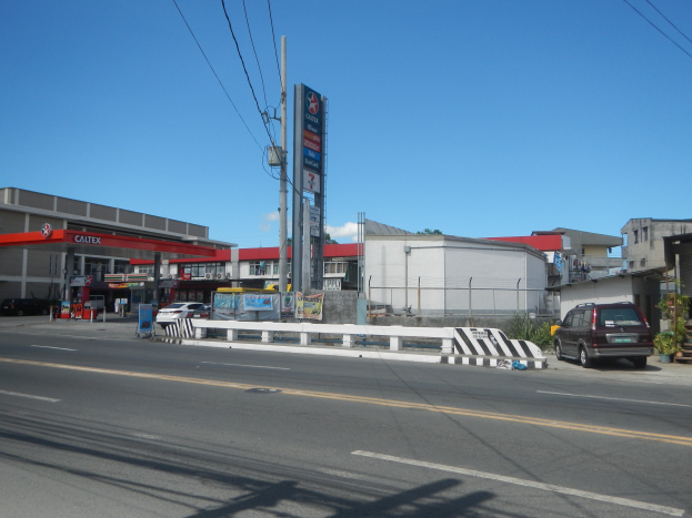 Tankstelle an einer Straßenkreuzung mit parkenden Autos, Gebäuden, Strommasten, Schildern, Geländern, Pflanzen und einem bewölkten Himmel im Hintergrund.