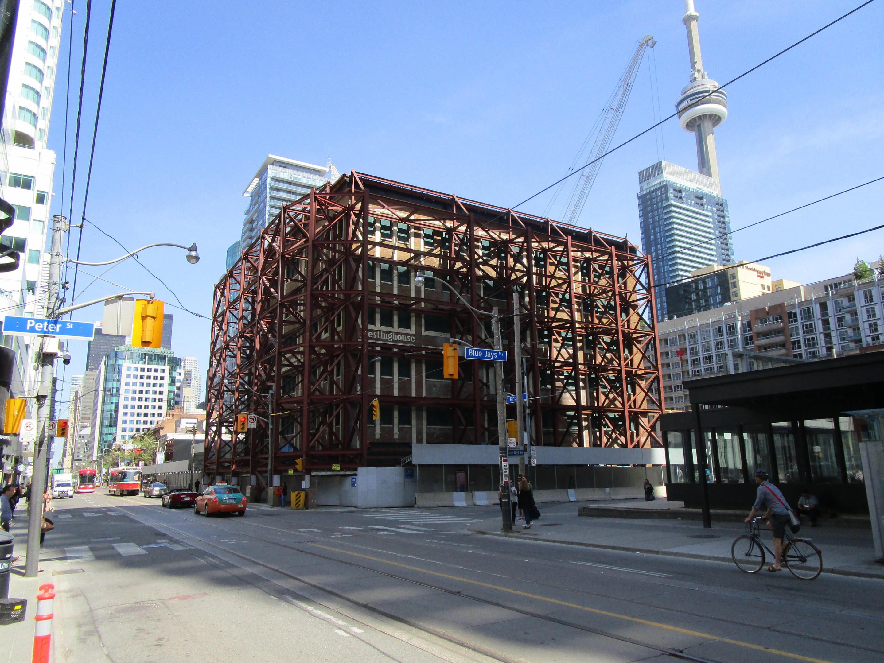 Eine belebte Stadtstraße mit einem großen Gebäude im Hintergrund, das eine Baustelle zeigt, mit Fahrzeugen, einem Radfahrer, Passanten, Ampeln, Pfählen, Schildern, einem Kran und Gerüsten.
