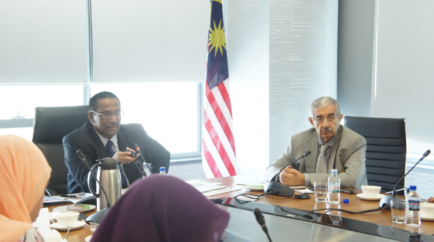 Eine Gruppe von Menschen, darunter Männer in Anzügen und zwei Frauen in Burkas, sitzt um einen Tisch mit Wasserflaschen, einem Mikrofon, Tassen und einem Teekessel, wobei im Hintergrund ein Fenster und eine Flagge zu sehen sind.