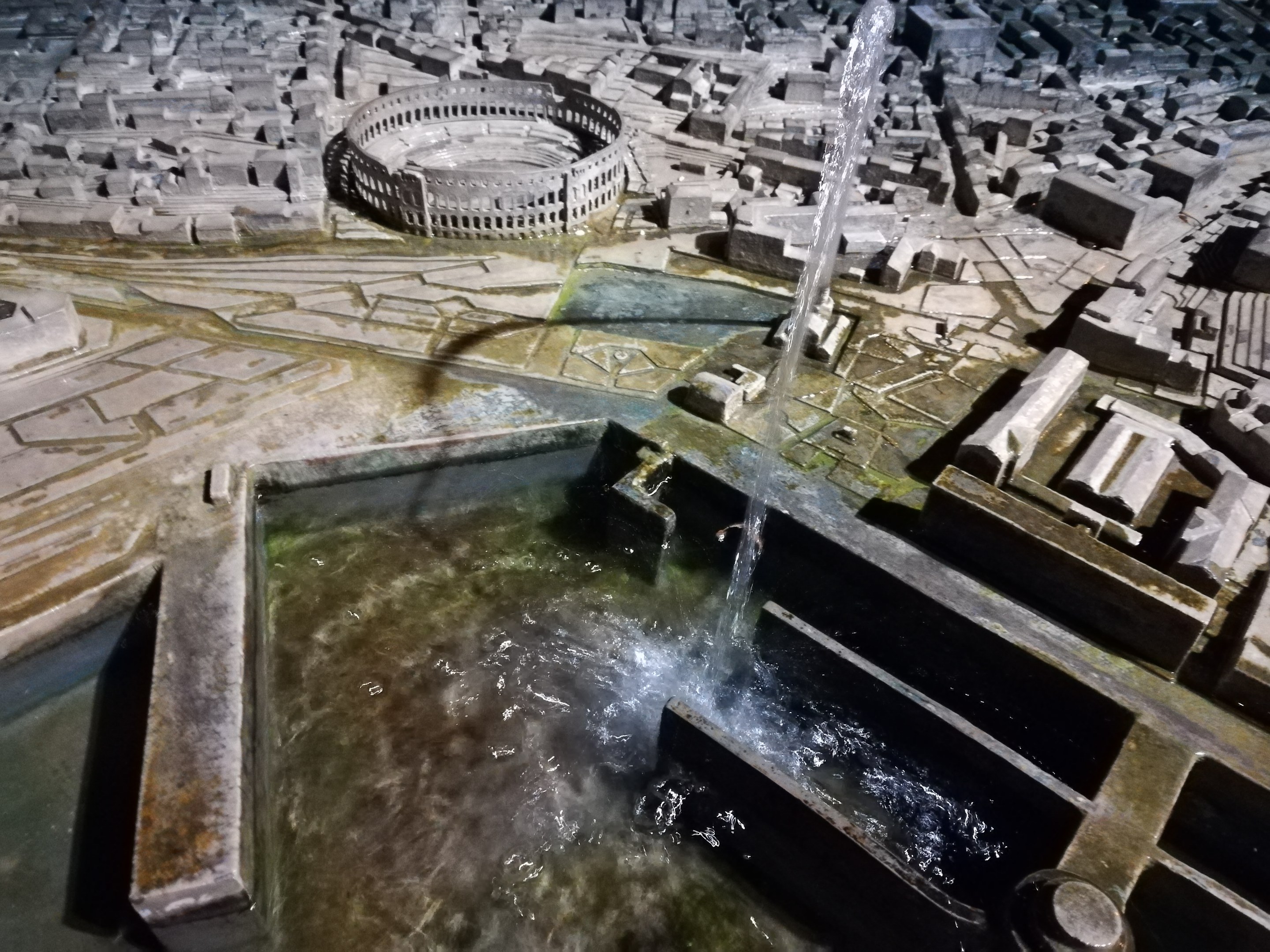 Ein Miniaturstadtmodell mit einem zentralen Brunnen mit Wasserfall, umgeben von Gebäuden und einer Wasserfläche.