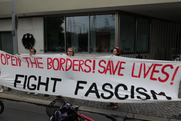 Eine Gruppe von Menschen mit einem Transparent mit der Aufschrift "Öffne die Grenzen, rette Leben, bekämpfe Faschismus" vor einem Gebäude mit Glasfenstern und einem Mast, mit einem Fahrrad und einer Tasche auf der Straße.