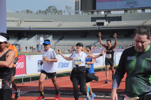 Gruppe von Menschen, die auf einer Stadionbahn laufen, mit Bannern, einer Anzeigetafel, einem Gebäude, Bäumen und einer bewölkten Himmel; einige Läufer tragen Turnschuhe und Hüte.