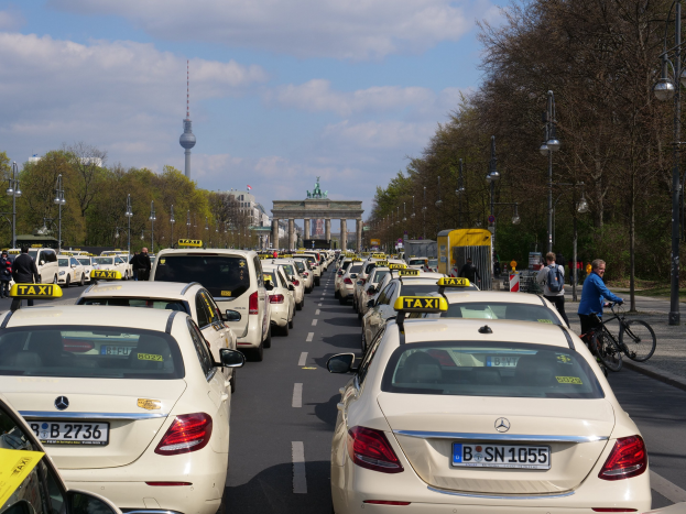 Eine lange Reihe von Taxis, die entlang einer belebten Straße in Berlin, Deutschland, geparkt sind, mit Fahrzeugen, Radfahrern und Fußgängern, flankiert von Laternenmasten und Bäumen, und Gebäuden, einem Bogen und einem Turm im Hintergrund unter einem bewölkten Himmel.