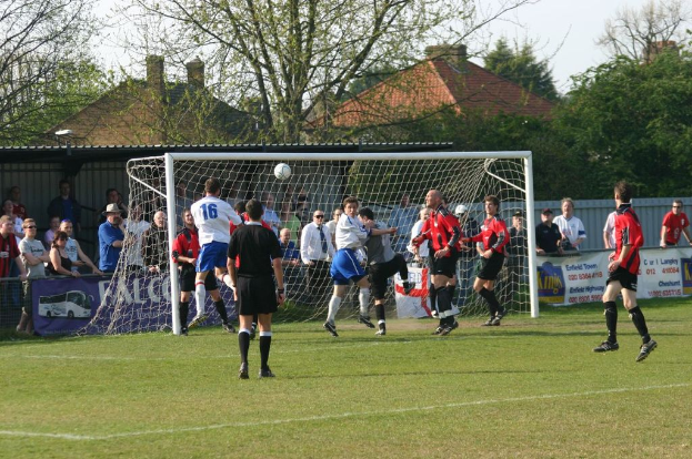 Fußballspieler sind in einem Spiel auf einem Feld mit einem Tornetz beteiligt, während Zuschauer dahinter stehen, mit Bäumen und Häusern im Hintergrund.