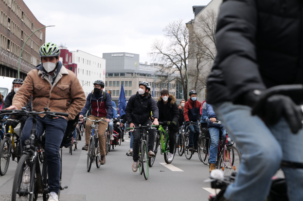 Eine Gruppe von Radfahrern in Helmen und Handschuhen fährt eine von Bäumen gesäumte Straße in Berlin entlang, mit Gebäuden, geparkten Fahrzeugen und Himmel im Hintergrund.
