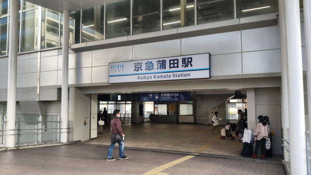 Menschen mit Taschen durch eine Bahnhofshalle gehen, eine Abfahrtsanzeige, Säulen, Geländer, Glaswände und Deckenlampen; ein Mann in der Mitte mit Schuhen.