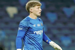 Ein junger Mann in einem blauen Torwarttrikot steht auf einem Fußballfeld und trägt ein blaues T-Shirt und weiße Handschuhe während eines Spiels.