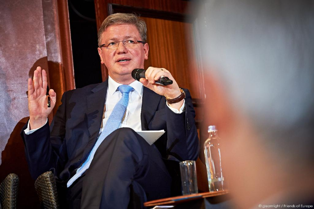 Ein Mann sitzt auf einem Stuhl neben einer hölzernen Tür, hält ein Mikrofon und einen Stift in der Hand, mit einem Glas, einer Wasserflasche und anderen Gegenständen auf dem Tisch daneben, und Text in der oberen rechten Ecke.