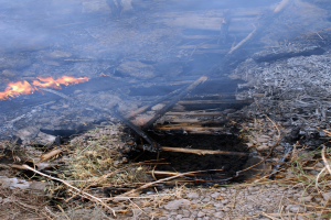 Ein Feuer brennt hell inmitten eines Schuttesfeldes, umgeben von Gras, Steinen und Holzstöcken, erhellt die Umgebung.