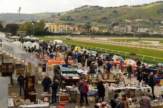Große Gruppe von Menschen, die einen Flohmarkt durchstöbern, mit Tischen, auf denen Gegenstände wie Foto Rahmen und Stühle ausgelegt sind, geparkte Fahrzeuge, Geländer, Stufen, Bäume, Gebäude, Laternenpfähle, Hügel und ein bewölkter Himmel im Hintergrund.