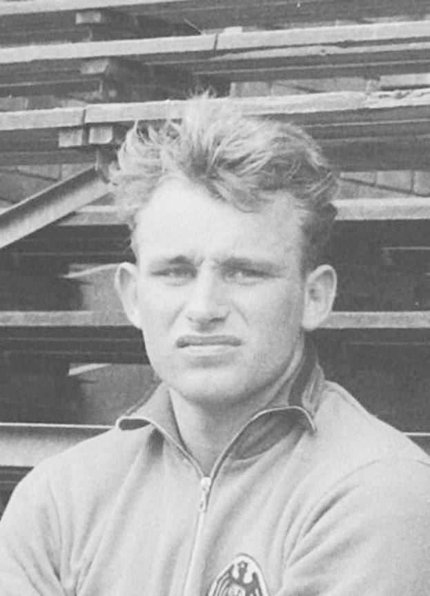 Schwarz-weißes Foto eines Mannes in einer Jacke vor gestapelten Holzplanken, der legendäre deutsche Fußballspieler soll es sein.