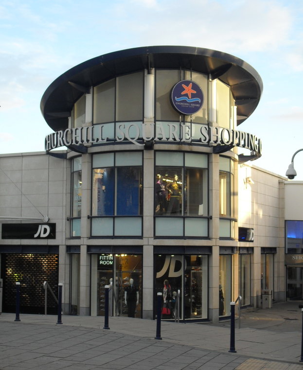 Eine belebte Straße mit einem großen Einkaufszentrum namens "Churchill Square Shopping" in der Mitte, umgeben von Straßenmasten und Lichtern, mit Schaufensterpuppen in Kleidern im Innern.