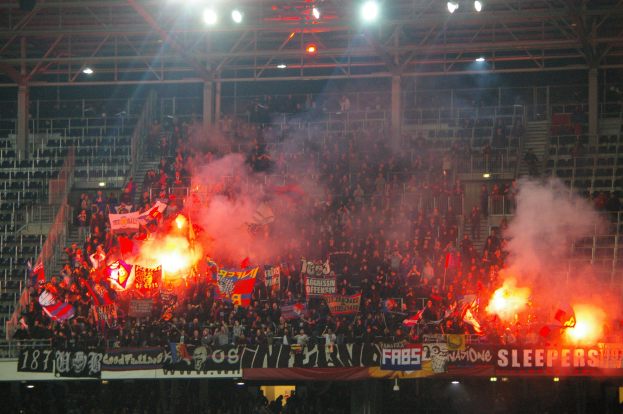 Eine große Menge Menschen in einem Stadion hält Fahnen und Banner, mit Leuchtraketen und Rauch, der aus ihnen aufsteigt, während Metallrahmen und Deckenleuchten darüber sichtbar sind.
