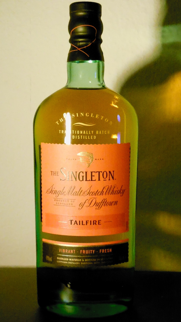 Eine tiefe bernsteinfarbene Flasche Singleton of Dufftown Tailfire Single Malt Scotch Whisky auf einem Tisch, mit einem schwarzen Etikett, das den Namen des Whiskys und ein Wappen mit einem Löwen und einer Burg zeigt.