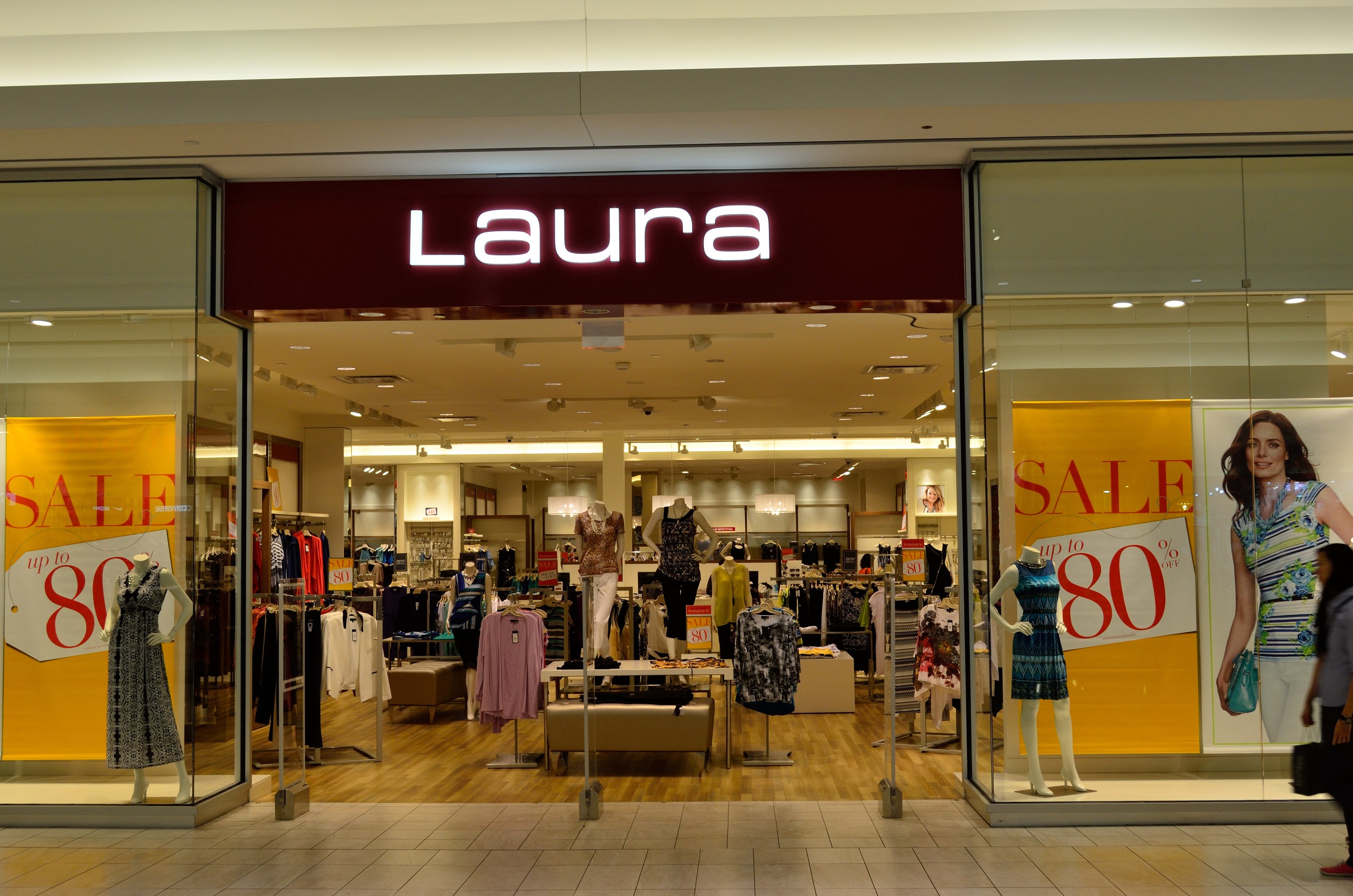 Ein Laura-Kleidungsgeschäft in einem Einkaufszentrum mit Schaufensterpuppen in Kleidern, hängenden Kleidern, Informationsschildern, Deckenlampen und einer Person mit einer Tasche.