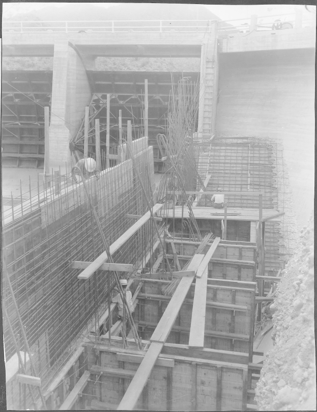 Ein Schwarz-Weiß-Foto einer Baustelle mit einer Brücke im Hintergrund, das Arbeiter, Stangen, hölzerne Gegenstände und andere Baustoffe zeigt, die über die Fläche verstreut sind, mit Text am unteren Rand des Bildes.