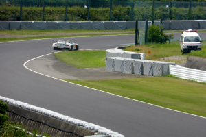 Ein Rennwagen fährt auf einer Rennstrecke mit einem Polizeiwagen im Hintergrund, umgeben von Gras, Pflanzen, Bäumen und Zaun, mit einem Fahrzeug auf der rechten Seite und Text am unteren Rand.